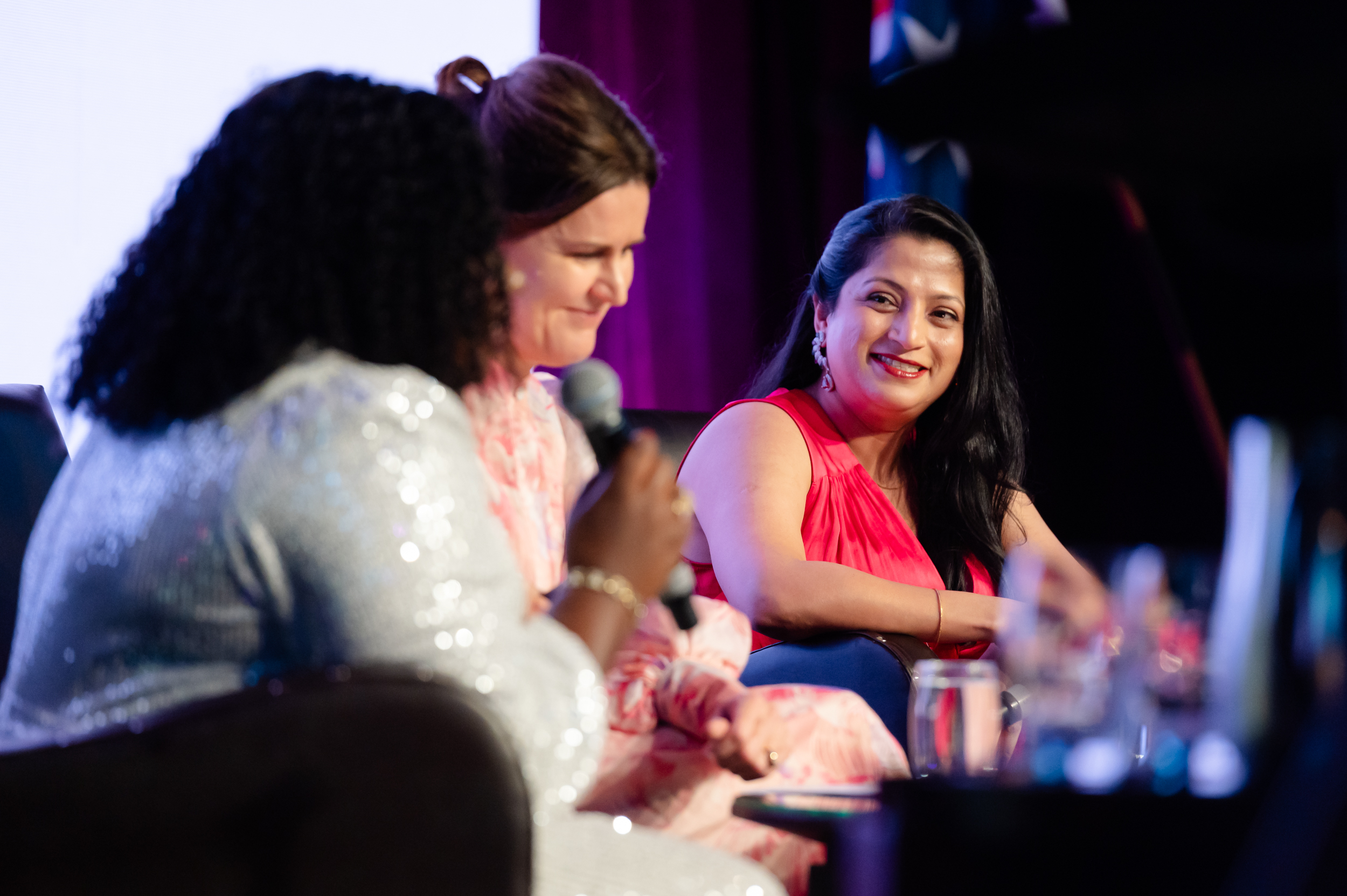 Three women sitting on sofa on stage talking, one holding a mic