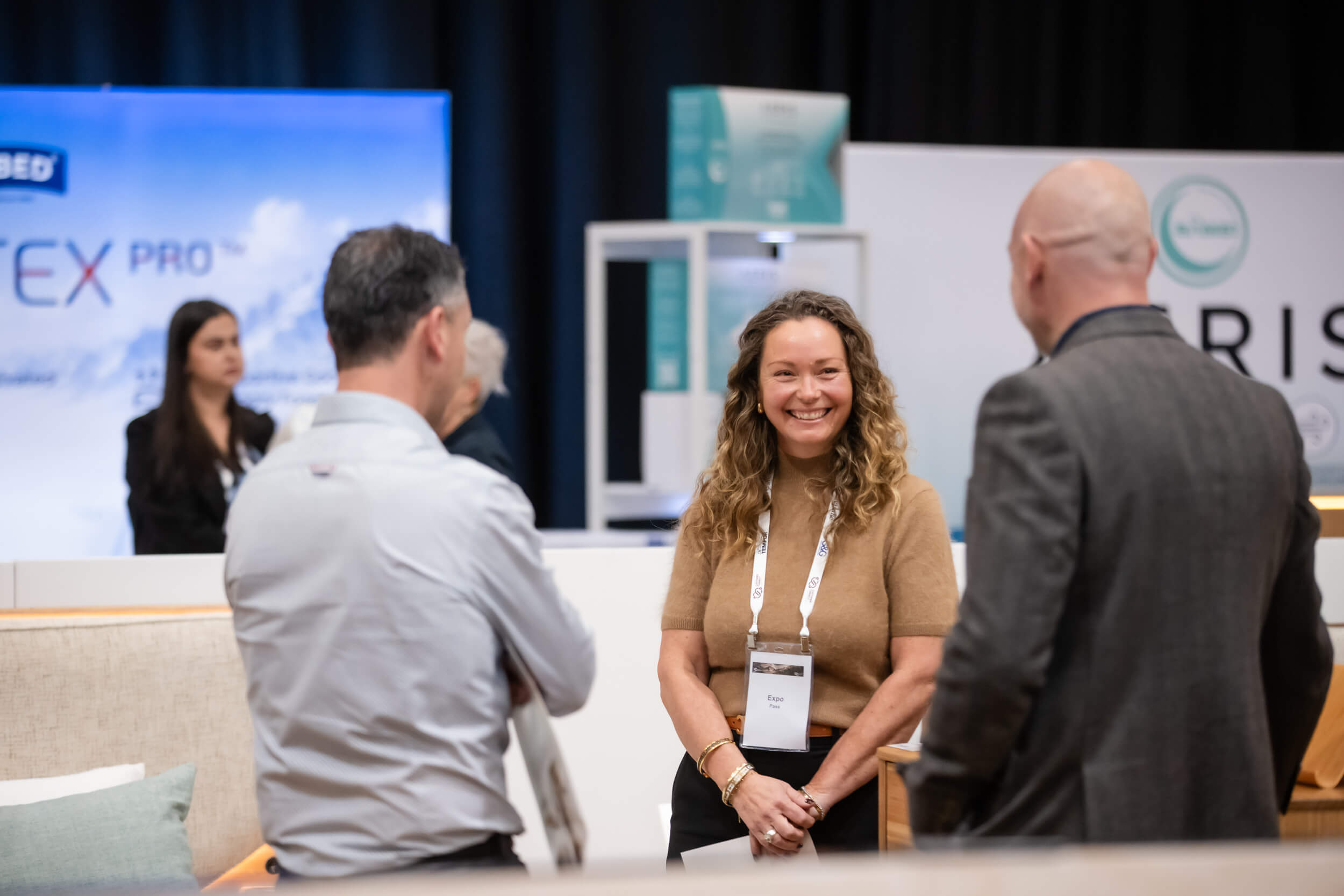 SELECTS-Snooze-Conference-01Sep26-002 A woman with curly hair smiles while engaging in conversation with two men at a conference. Branding displays are visible in the background.