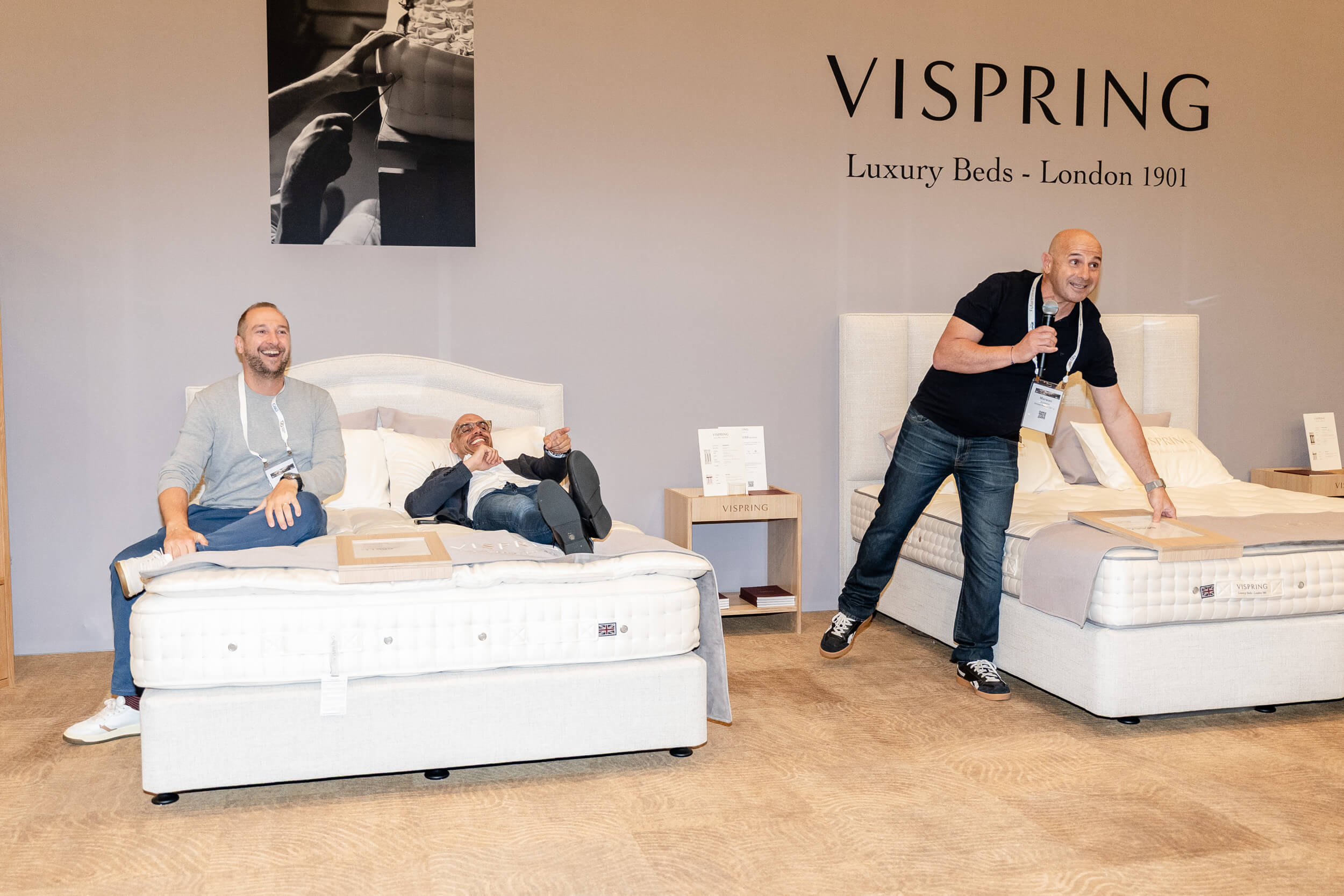SELECTS-Welcome-Dinner-01Sep26-015 Two men sitting on a luxury Vispring bed, with a third man standing and speaking, showcasing the beds at a display.